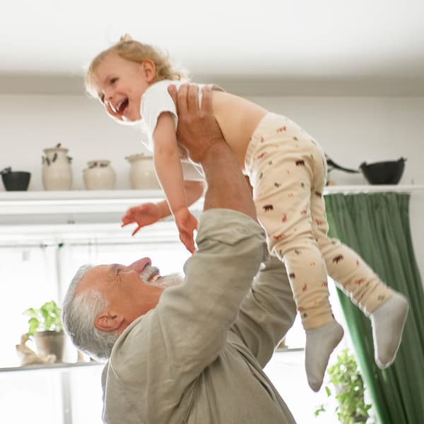 A smiling person with gray hair and beard uses both arms to hold above his head a smiling child, beneath a white ceiling.