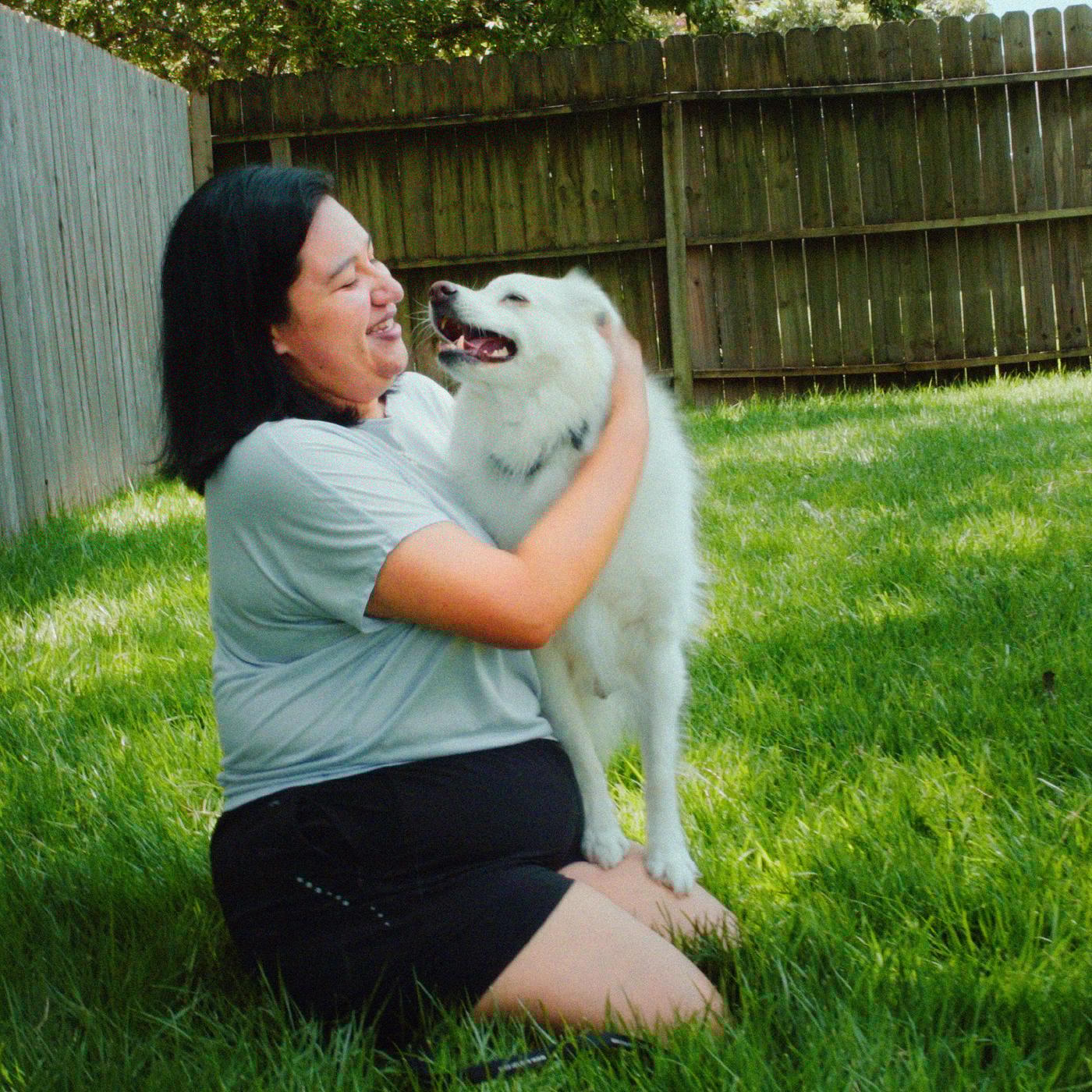 A smiling woman in a t-shirt and shorts is playing with a white dog on green grass.