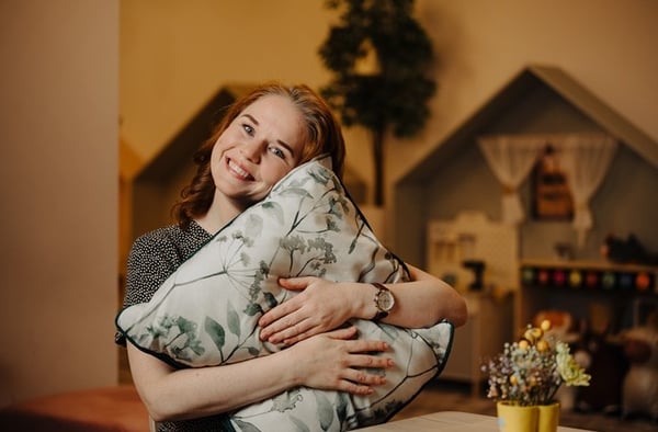 A smiling woman is leaning against a table indoors, with a cozy interior, plants, and decorations visible in the background.