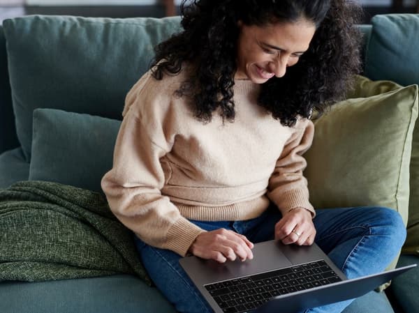 A smiling woman is sitting on a sofa with an open laptop on her lap