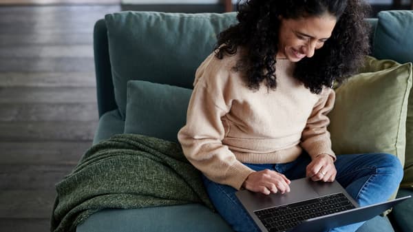 A smiling woman is sitting on a sofa with an open laptop on her lap