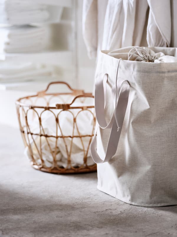 A SNIDAD basket holding laundry and a PURRPINGLA laundry bag stand on a floor in front of some shelves holding folded towels.