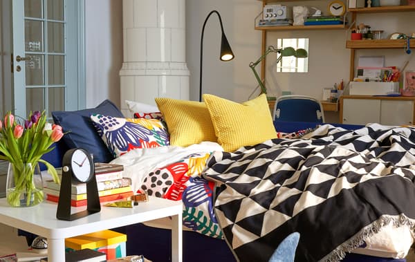 A sofa bed made up as a bed in a living and work space with a black floor lamp and a white side table.