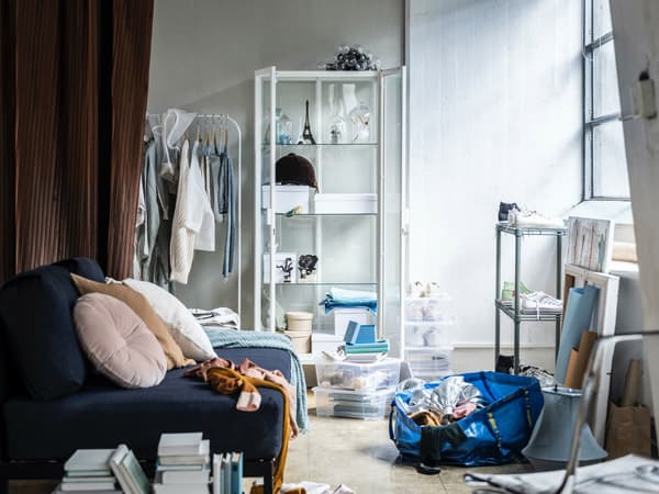 A sofa in a cluttered room with MILSBO cabinet standing at the back. A rack of clothes stands next to it.