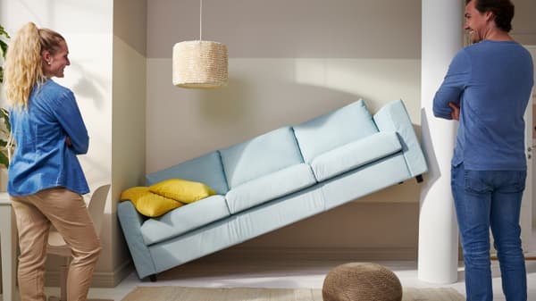 A sofa is wedged sideways between two walls. A man and a woman are looking at it, laughing because it doesn’t fit in the spot they’d planned.