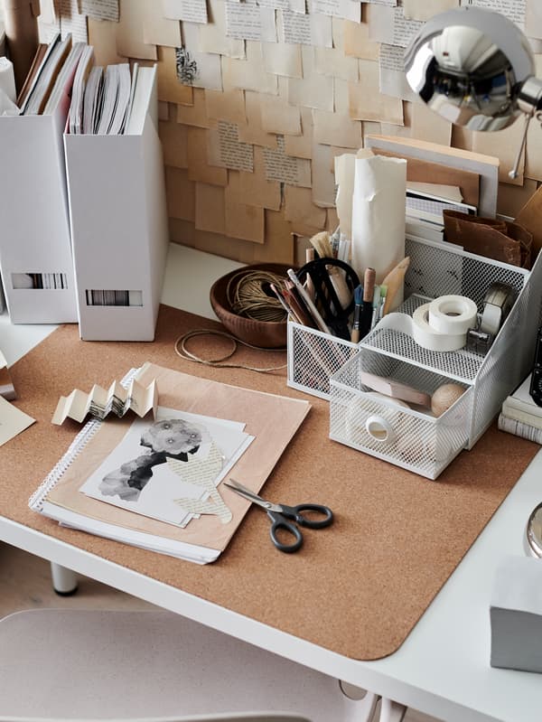 A somewhat cluttered workspace with a DRÖNJÖNS desk organiser, a SUSIG cork desk pad and a FORSÅ nickel-plated work lamp.
