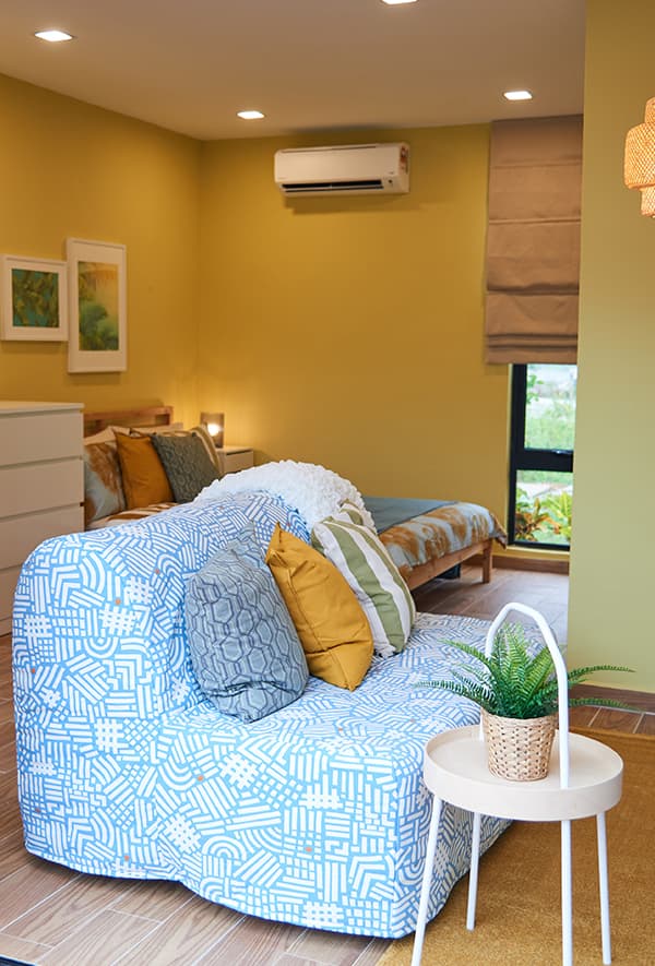 A sova with blue and white decorative sofa cover and a side table with a potted plant on top in a yellow room.