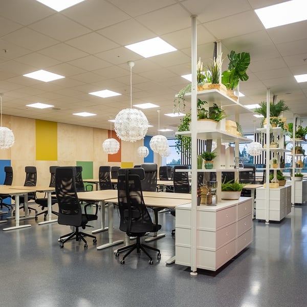 A spacious open-plan office features rows of large shared desks with black ergonomic chairs on wheels. The ceiling is fitted with bright square LED panels, and several spherical pendant lights made of intricate white floral patterns hang above the workstations. The back wall displays vertical color blocks in yellow, green, orange, and blue on a light wood paneling. White shelving units with drawers and decorative plants divide the space, adding greenery and a welcoming atmosphere. The overall design is modern, airy, and collaborative.