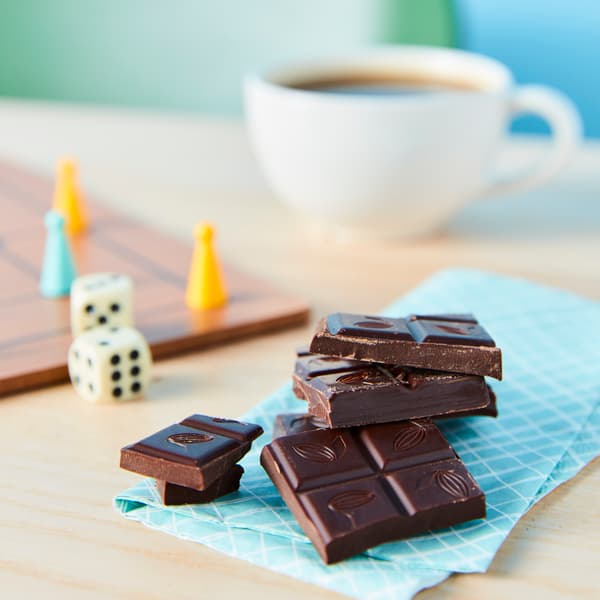 A stack of CHOKLAD MÖRK dark chocolate on a wooden surface next to a cup of coffee and a set of dice.