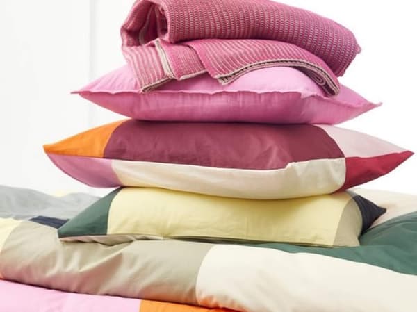 A stack of colorful pillows, a comforter and a knitted throw blanket. The bottom pillow is yellow, followed by a green, pink, and an orange pillow on top.