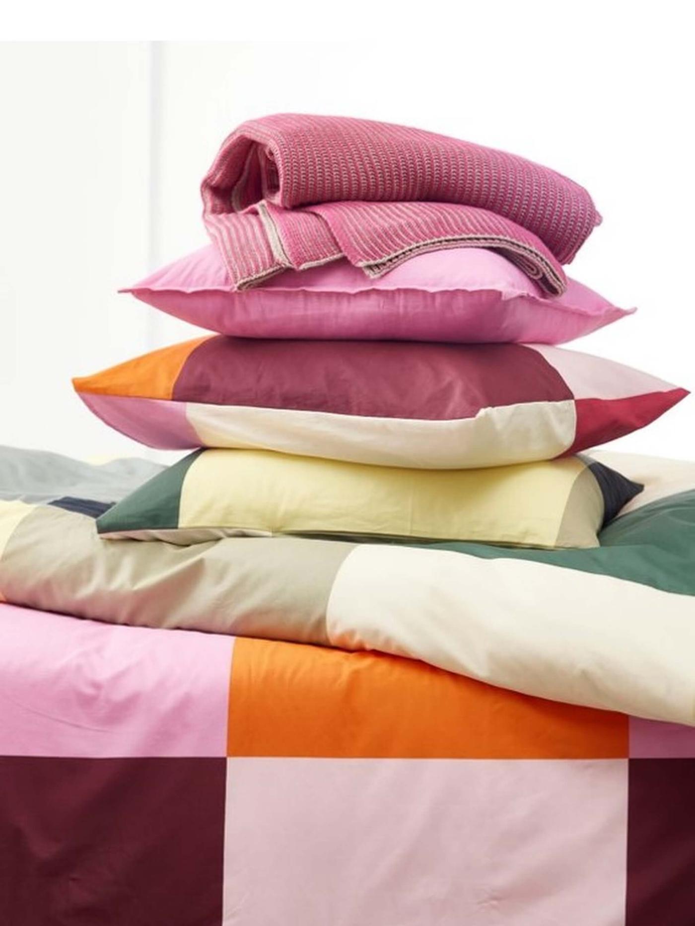 A stack of colorful pillows, a comforter and a knitted throw blanket. The bottom pillow is yellow, followed by a green, pink, and an orange pillow on top. 