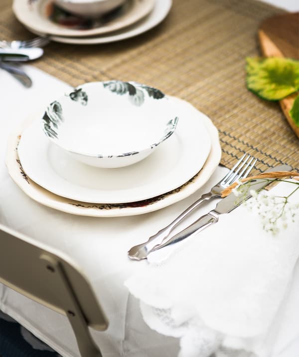 A stack of crockery and cutlery set on a seagrass table runner and white table cloth.