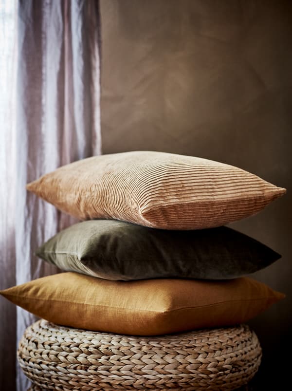 A stack of cushions, including ones in ÅSVEIG and VIGDIS covers, in earthy shades and resting on a woven banana-fibre stool.