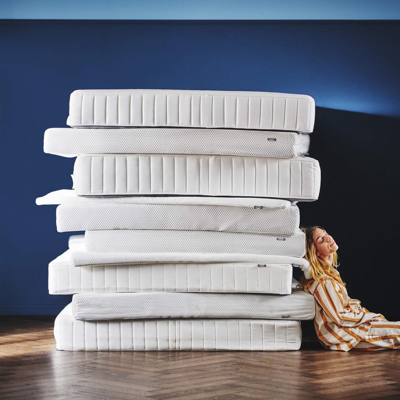 A stack of eight different white mattresses of varying thicknesses and designs, neatly piled on top of each other and a woman leaning against them.