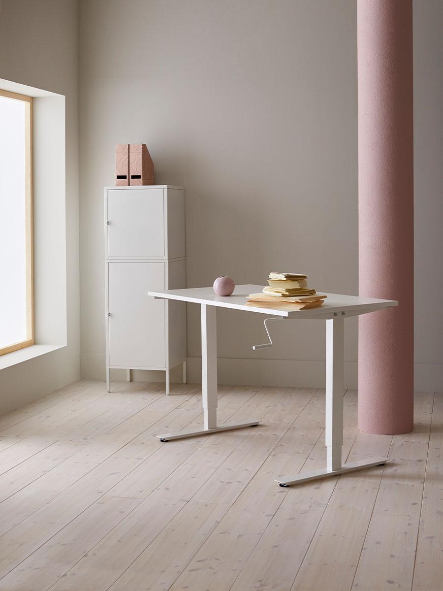 a stack of papers on a white desk in the middle of a bare room in front of a white cabinet with two magazine files on top