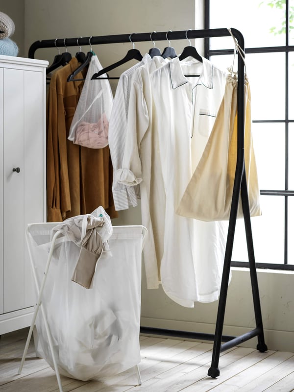 A standing clothes rack in black with clothes hanging on hangers in front of a window.