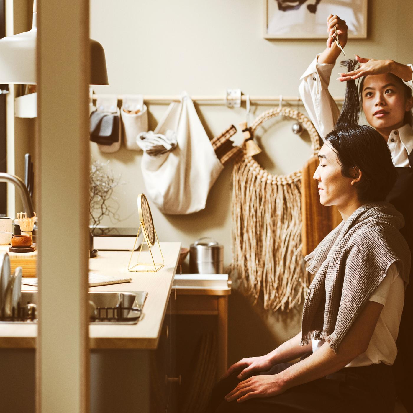 A standing person cutting the hair of a seated person in a kitchen with an ash effect EKBACKEN worktop.