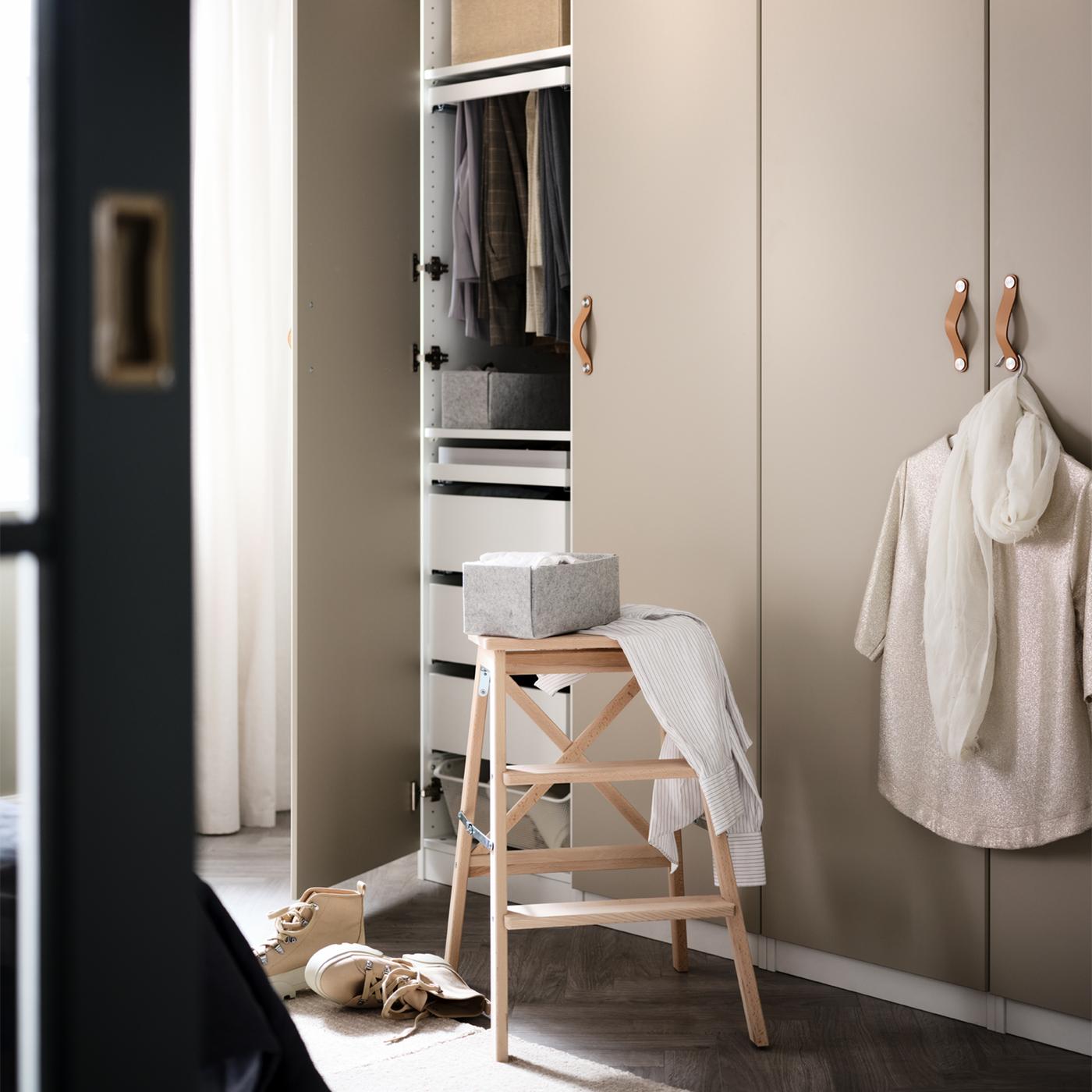 A step stool in front of an open door of a PAX/REINSVOLL wardrobe combination, revealing hanging clothes and drawers inside.