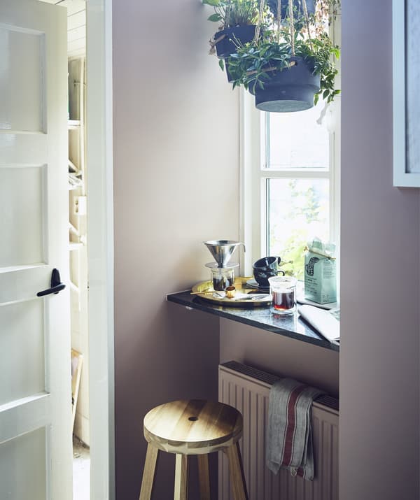 A stool and ledge in front of a large window with hanging plants and coffee-making equipment.