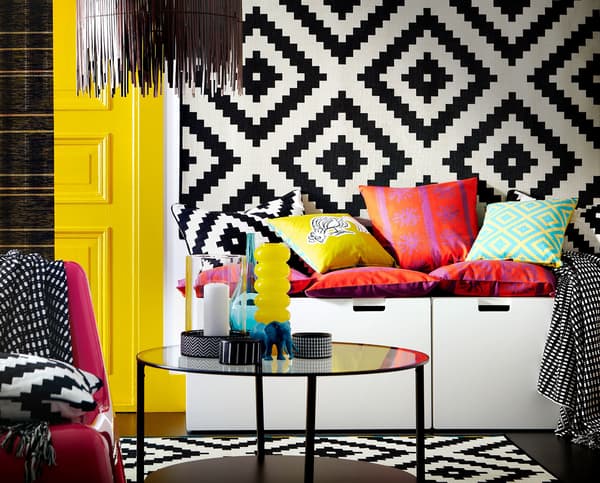 A storage bench with cushions on top for seating and a patterned rug hanging on a wall.