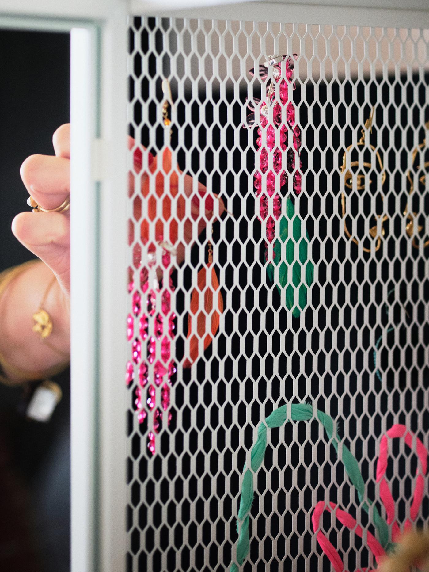 A storage unit with a vertical, white metal mesh surface. A woman’s hand is hanging several earrings in the mesh holes.