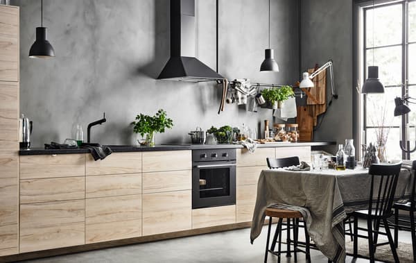 A straight-line kitchen with a row of base cabinets with ash fronts against a grey, concrete looking wall  and a dinng table with a linen table cloth