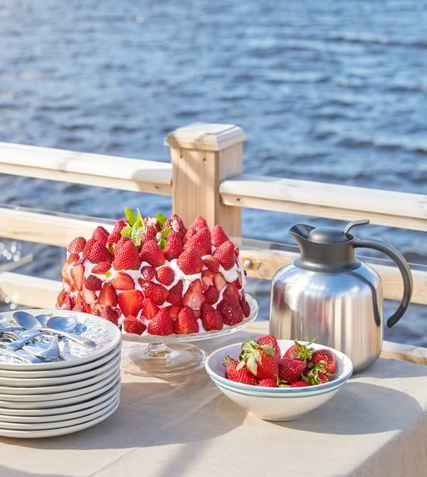 A strawberry and cream cake with a bowl of strawberries and a stack of plates.