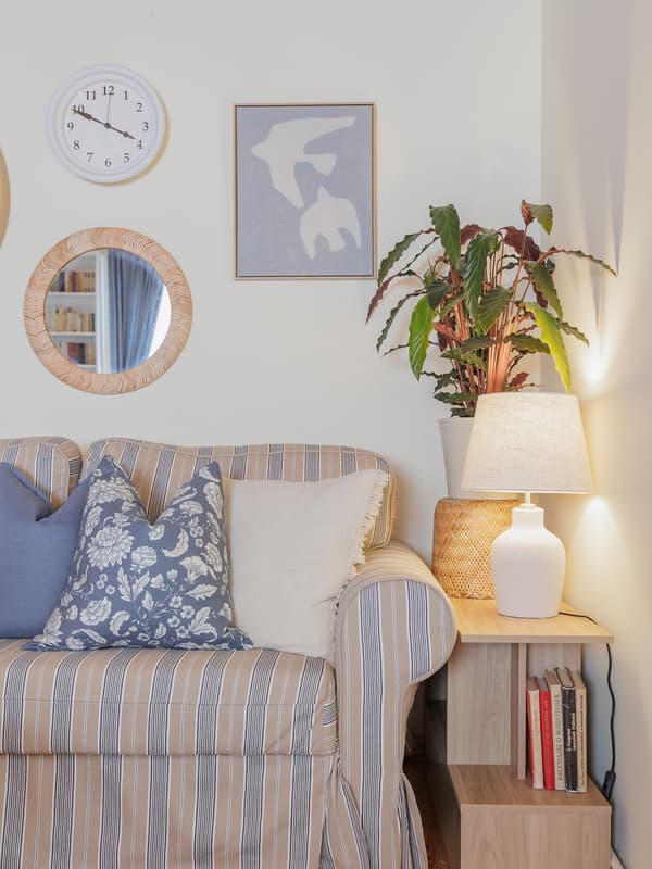 A striped sofa-bed with blue and floral cushions sits beneath a gallery wall with round mirrors and framed art. A wooden side table with a ceramic lamp and potted plant completes the look.