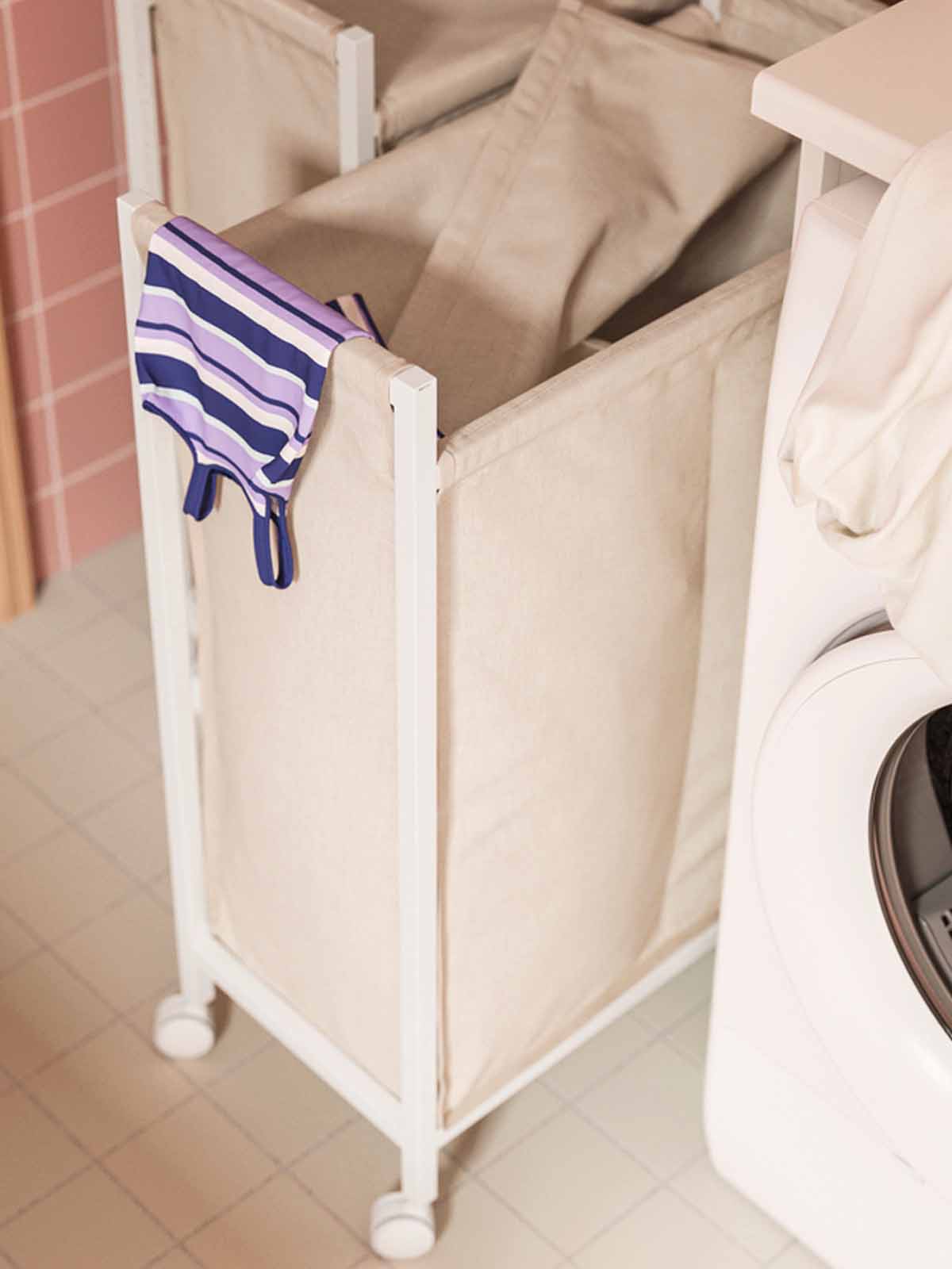 A striped t-shirt peeking out of a white ENHET laundry trolley with castors besides a washing machine at the laundry room.