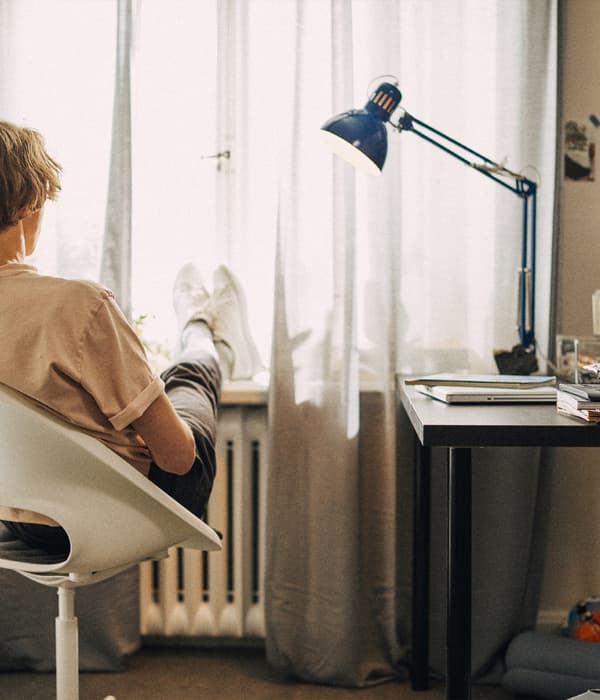 A student seated in a white chair relaxing next to a desk with a blue work lamp on it.