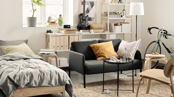 A studio room with a bed, GLOSTAD sofa and an open shelving unit against the wall. A small table is in front of the sofa.