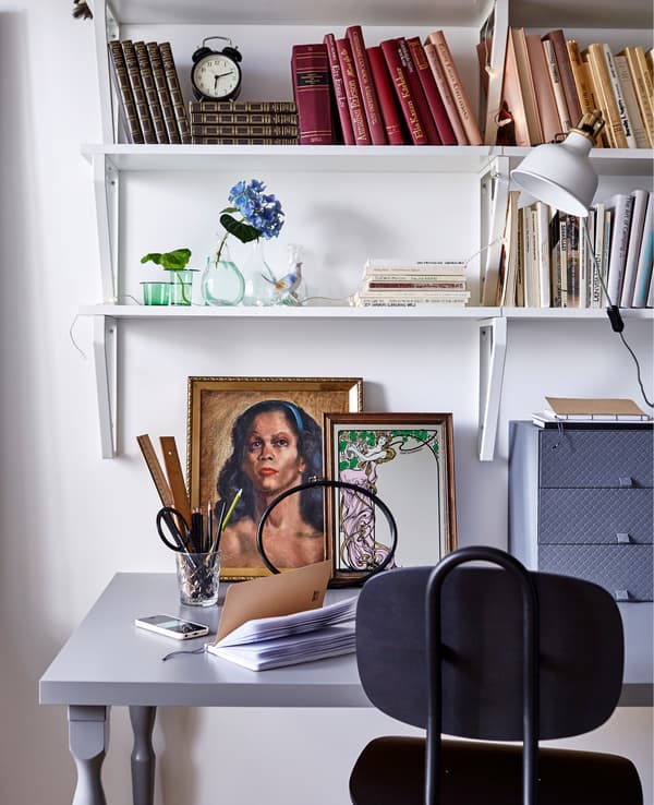A study space in a white bedroom, featuring a black office chair, a grey desk, some artwork and wall shelves containing books.