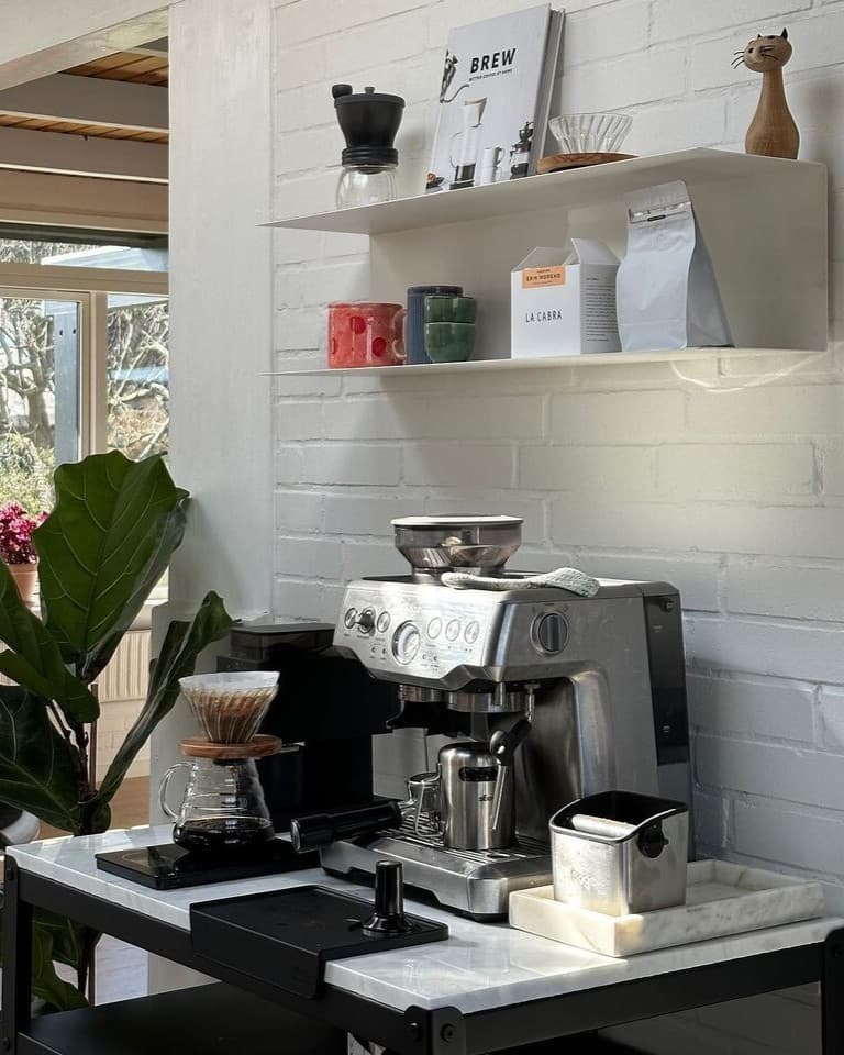 A stylish coffee corner with a stainless steel espresso machine and pour-over coffee maker on a mobile trolley, a white brick wall with a floating shelf filled with coffee cups, coffee bags, and accessories, next to a large green houseplant by the window.
