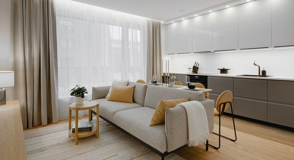 A stylish open-concept living room and kitchen area featuring a light gray sofa with mustard yellow accent pillows and a fringed throw. Behind the sofa is a modern dining setup with a wooden bar-height table and matching chairs. The kitchen has sleek, minimalist cabinetry in matte gray with integrated lighting and built-in appliances. Sheer white curtains diffuse natural light from the large windows, complemented by beige drapes. A small side table with a potted plant and books sits next to the sofa on a light textured rug over wooden flooring, creating a cozy, contemporary atmosphere.