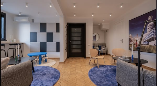 A stylish waiting area featuring light wooden flooring, blue circular rugs, modern gray sofas, black and wooden chairs, and small round coffee tables with minimalistic decor. The room includes a checkered acoustic panel on one wall, a small kids' table with stools, and a cozy reading nook with armchairs and a round mirror in the back. A large painting with a space shuttle and buildings decorates the right wall. Ceiling spotlights provide ample lighting throughout the space.