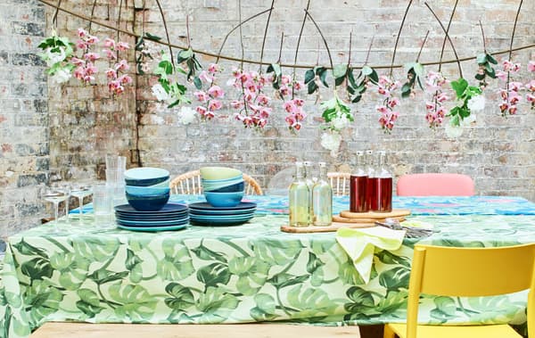 A summer party table with bright tablecloths, piles of blue crockery, mixed chairs and a garland of artificial flowers.