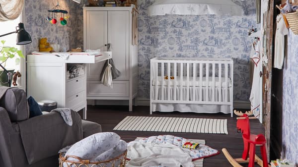 A SUNDVIK cot, wardrobe and changing table stand in a traditional children’s room with blue and white wallpaper.