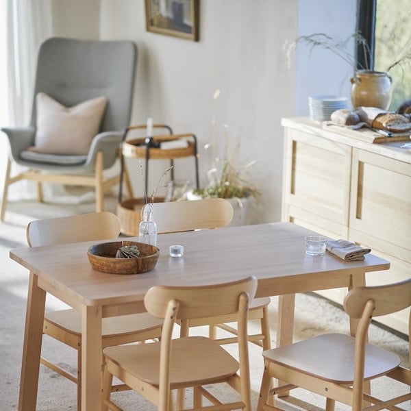 A sunlit dining area with a natural, rustic charm. A light wood dining table is surrounded by four matching chairs. On the table are simple accessories: a wooden bowl with dried botanicals, a clear glass vase with dried stems, a cloth napkin, and two drinking glasses. In the background, a light gray high-back armchair with a beige cushion sits near a small bar cart and a bundle of dried grasses. A sideboard against the wall holds plates, a ceramic jug, and fresh bread on a tray. The soft sunlight filtering through the window enhances the cozy, Scandinavian-inspired atmosphere.