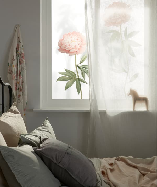 A sunlit window that’s partly covered by a sheer curtain and decorated with large floral deco stickers, in a bedroom setting.