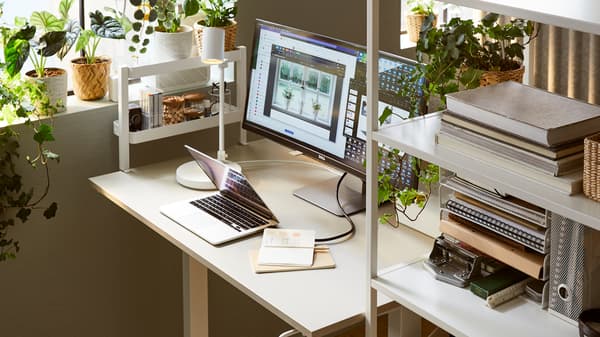 A sunlit workspace with a TROTTEN sit-stand desk between a white TROTTEN sliding-door cabinet and a window with plants.