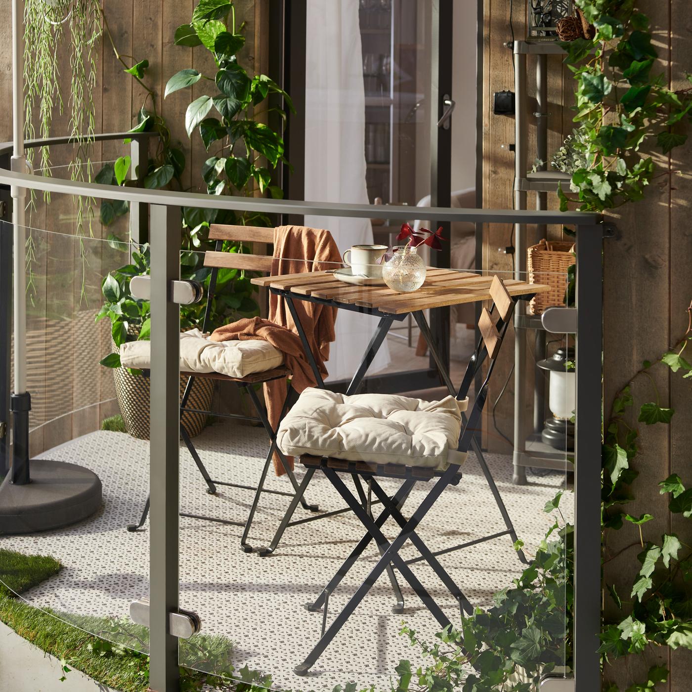 A sunny balcony featuring a TÄRNÖ table and two TÄRNÖ chairs with chair cushions on them, light grey floor decking and plants.