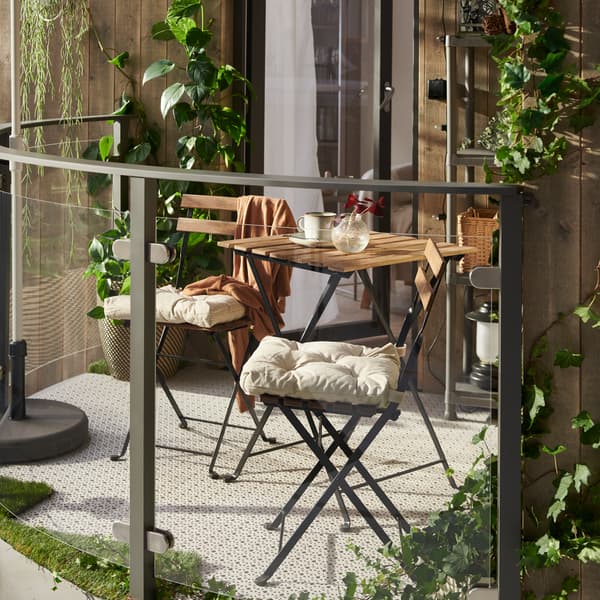 A sunny balcony featuring a TÄRNÖ table and two TÄRNÖ chairs with chair cushions on them, light grey floor decking and plants.