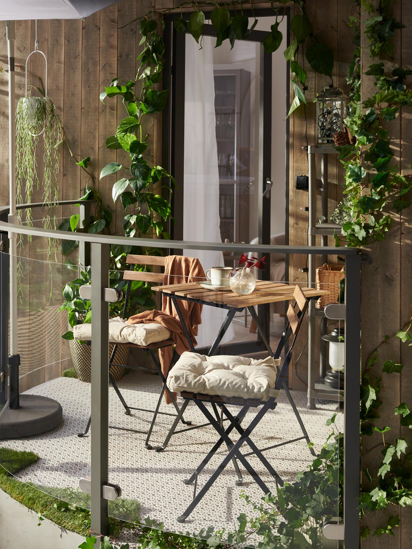 A sunny balcony featuring a TÄRNÖ table and two TÄRNÖ chairs with chair cushions on them, light grey floor decking and plants.