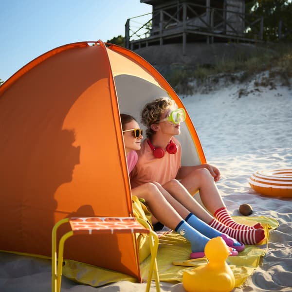 A sunny beach scene featuring a bright orange STRANDÖN sun shelter providing shade for two children on the sand.