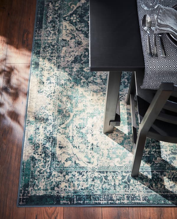 A table and a chair stand on a green/oriental-patterned rug. Cutlery and a black/white table-runner are on the table.