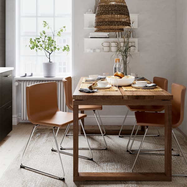 A table in brown-stained oak veneer, four chairs in golden-brown leather, a pendant lamp in seagrass and a beige rug.