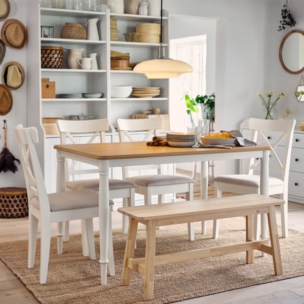 A table in oak/white, four white chairs and a bench in birch stand on a jute rug. Two high cabinets are in the background.