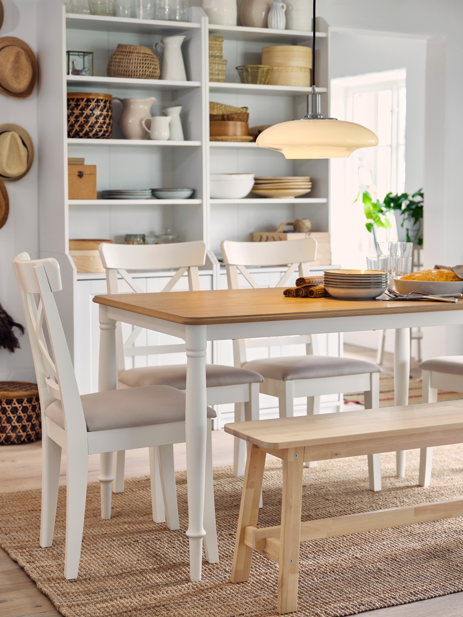 A table in oak/white, four white chairs and a bench in birch stand on a jute rug. Two high cabinets are in the background.