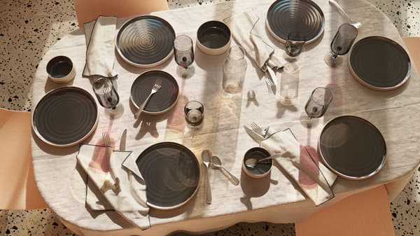 A table is set with OMBONAD dark grey 
plates and bowls, grey glasses and linen tablecloth; shadows play on the table.