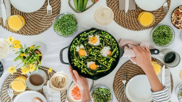 A table set for brunch, featuring a skillet with eggs, asparagus, and peas, surrounded by plates, glasses of orange juice, and fresh greens. A hand serves the dish.